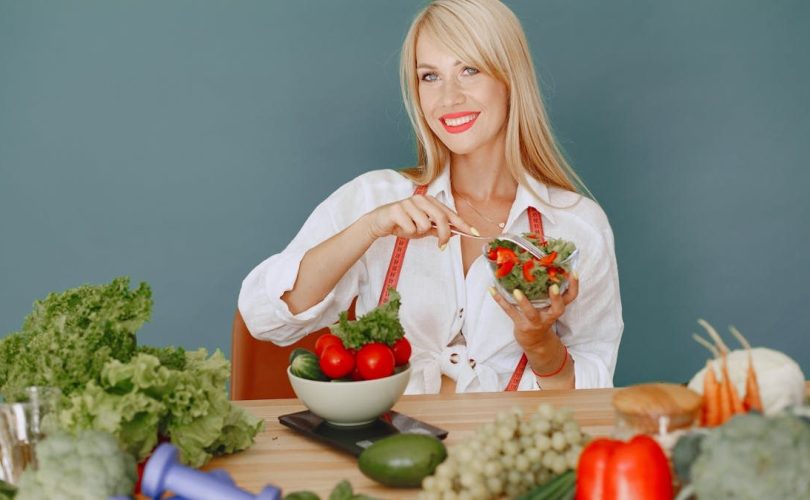 Blonde woman enjoying a fresh salad surrounded by vegetables, promoting healthy eating.