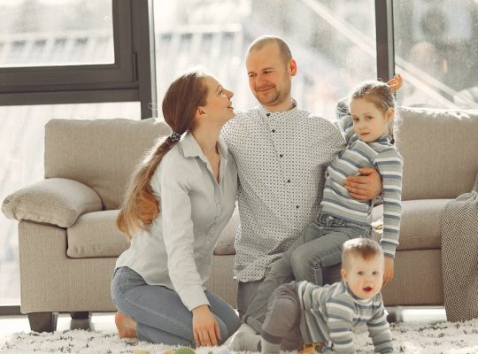 Cheerful mother and father smiling at each other while little cute boy crawling on floor and preschool girl in striped turtleneck raising arm and looking at camera