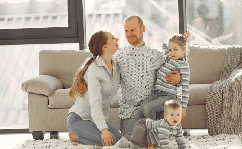 Cheerful mother and father smiling at each other while little cute boy crawling on floor and preschool girl in striped turtleneck raising arm and looking at camera